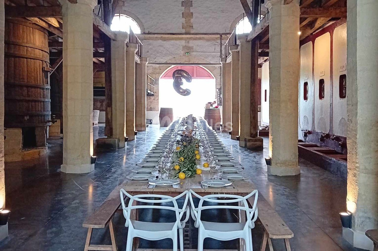 Grande table dressée avec vaisselle et décoration florale dans une salle voûtée avec tonneaux et cuves en arrière-plan.