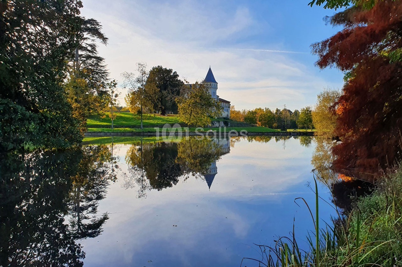Vue d'un château entouré d'arbres, reflété dans un étang calme sous un ciel dégagé.