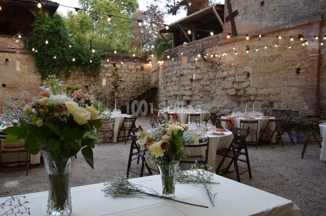 Décoration florale sur des tables dans une cour en pierre éclairée par des guirlandes lumineuses.
