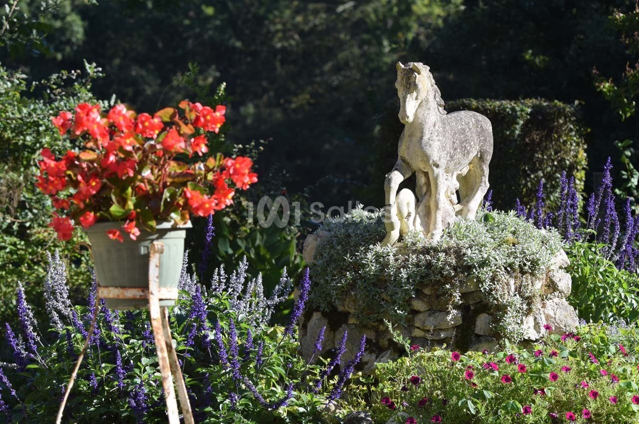 Statue de cheval avec des poulains sur un socle fleuri, entourée de plantes et de fleurs colorées dans un jardin.