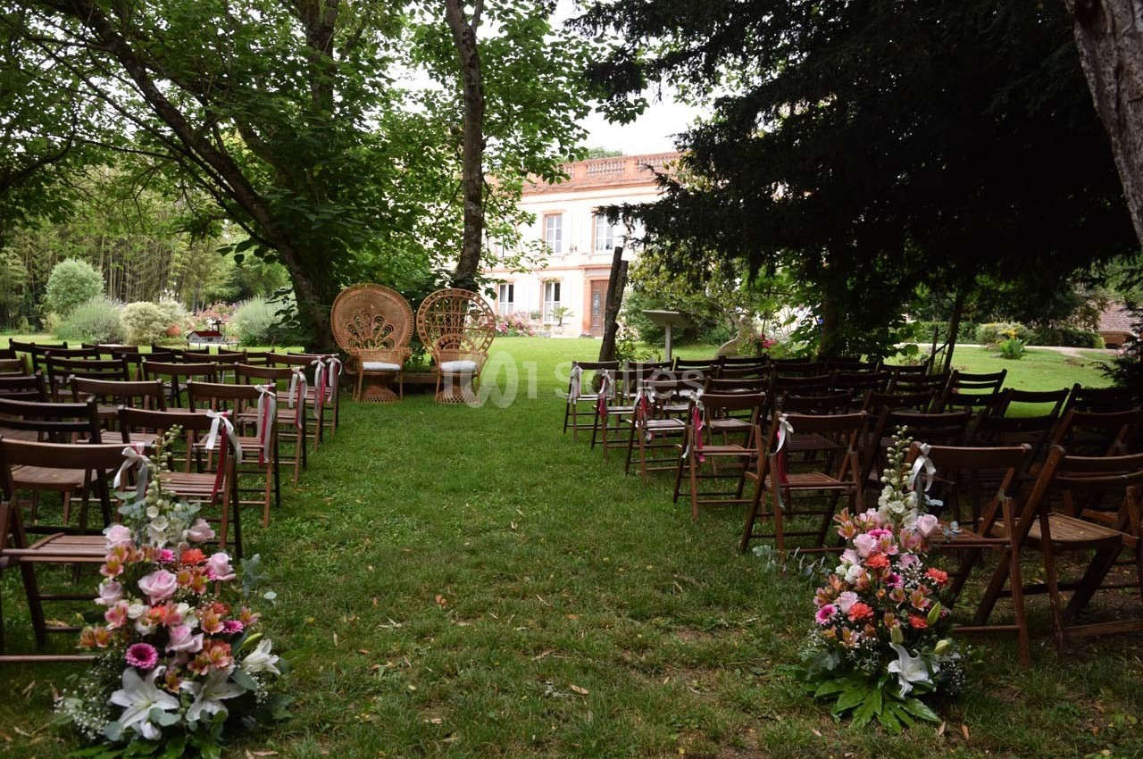 Chaises en bois disposées en allées sur une pelouse, décorées de fleurs, face à deux fauteuils en osier sous des arbres.