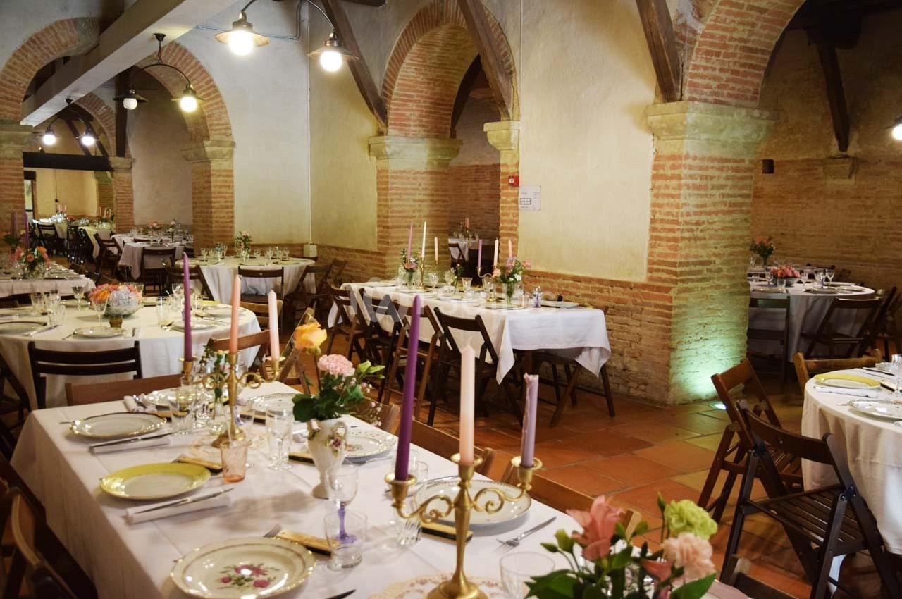 Salle voûtée avec tables dressées pour un repas, décorée de chandeliers, fleurs et vaisselle élégante.
