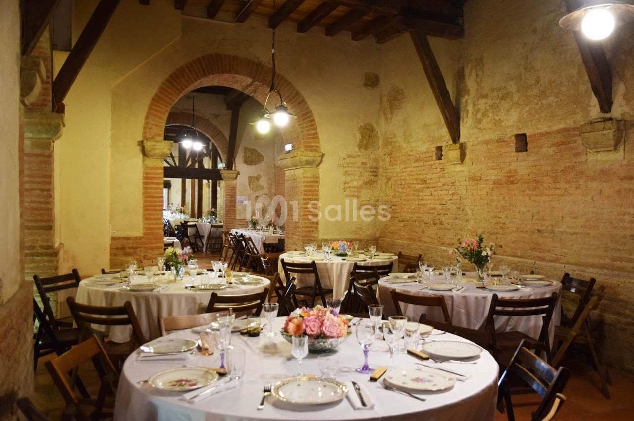 Salle rustique avec murs en briques, tables rondes dressées avec nappes blanches, vaisselle élégante et décor floral.