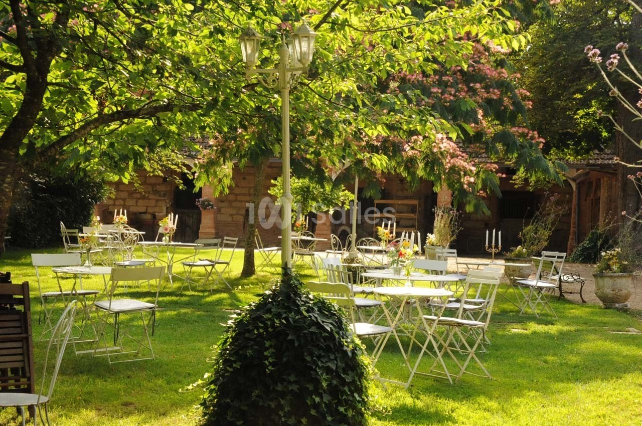 Chaises et tables blanches disposées dans un jardin verdoyant, entouré d'arbres et éclairé par un lampadaire.