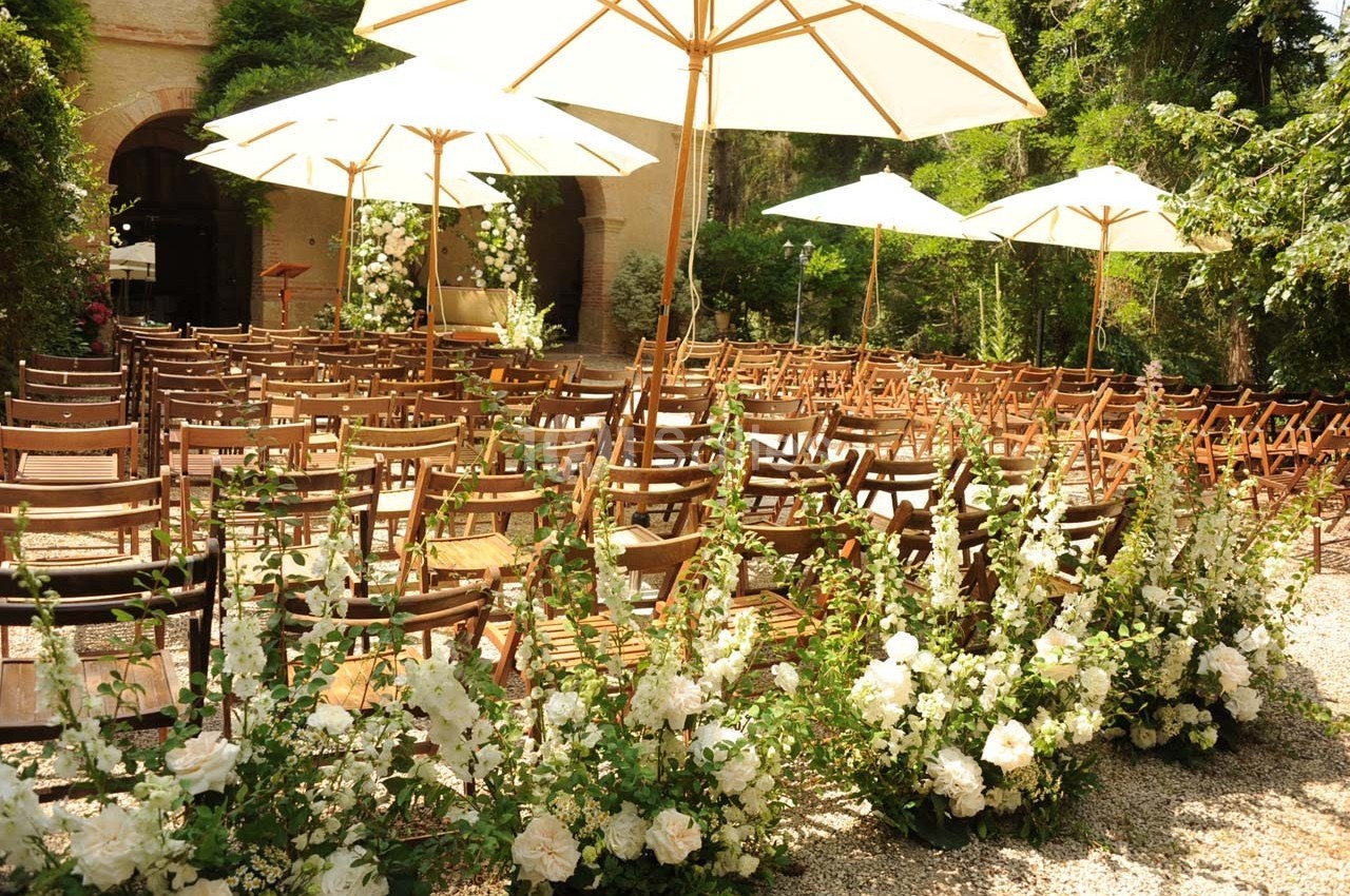 Chaises en bois alignées sous des parasols blancs, entourées de fleurs blanches, dans un jardin ensoleillé.