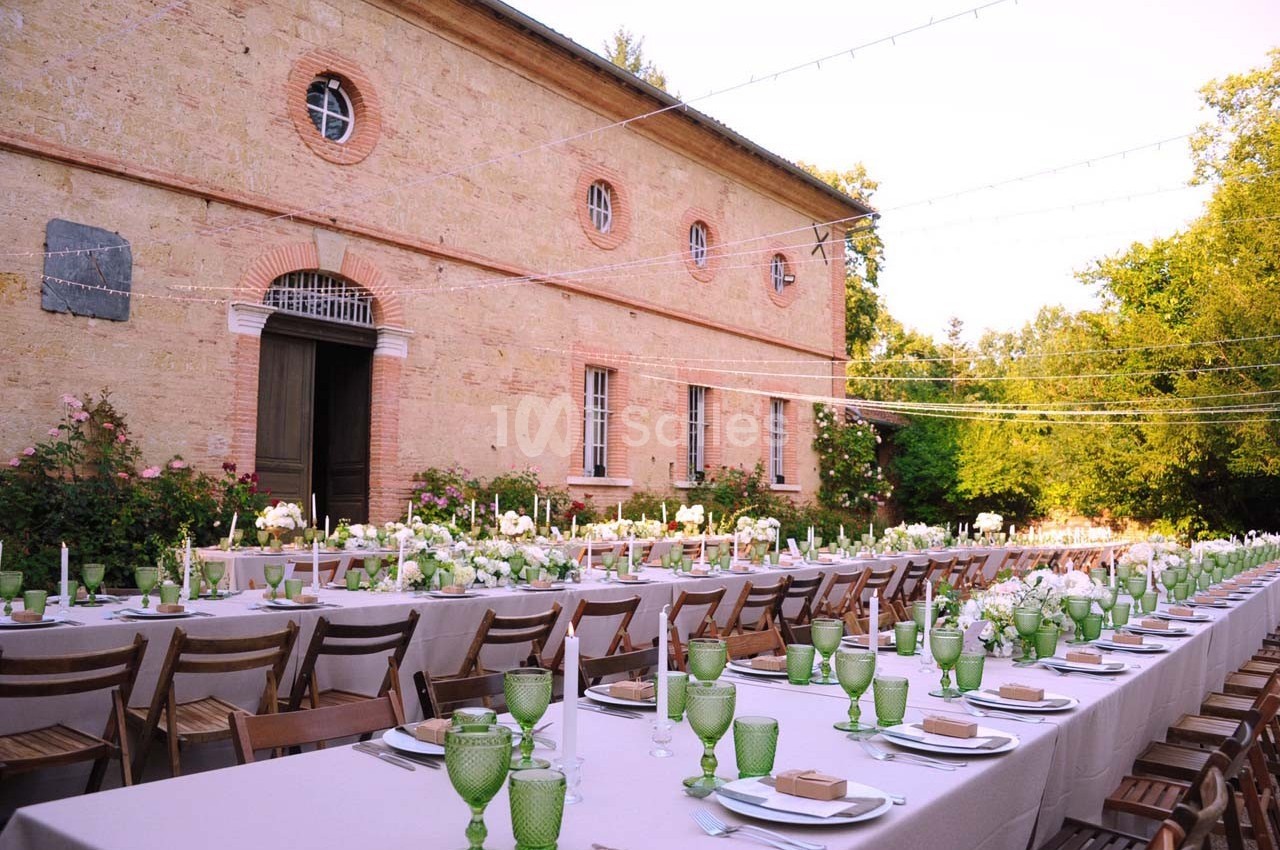 Grande table extérieure dressée avec vaisselle verte et décor floral, devant un bâtiment en pierre et un jardin verdoyant.