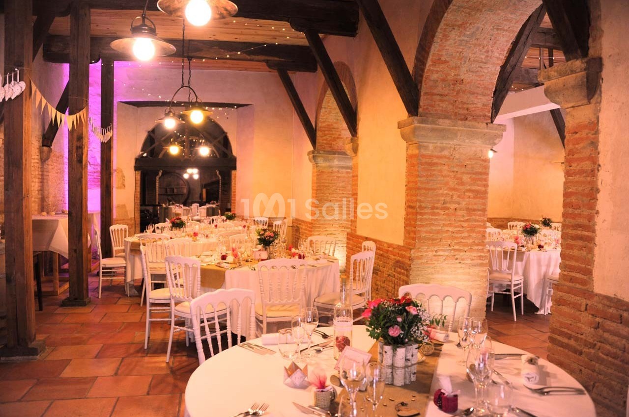 Salle de réception avec tables rondes décorées, chaises blanches et éclairage chaleureux sous des arches en briques.