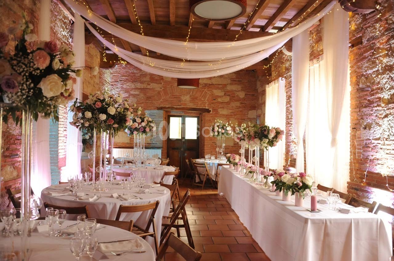 Salle de réception décorée pour un mariage avec tables dressées, fleurs et guirlandes lumineuses suspendues.