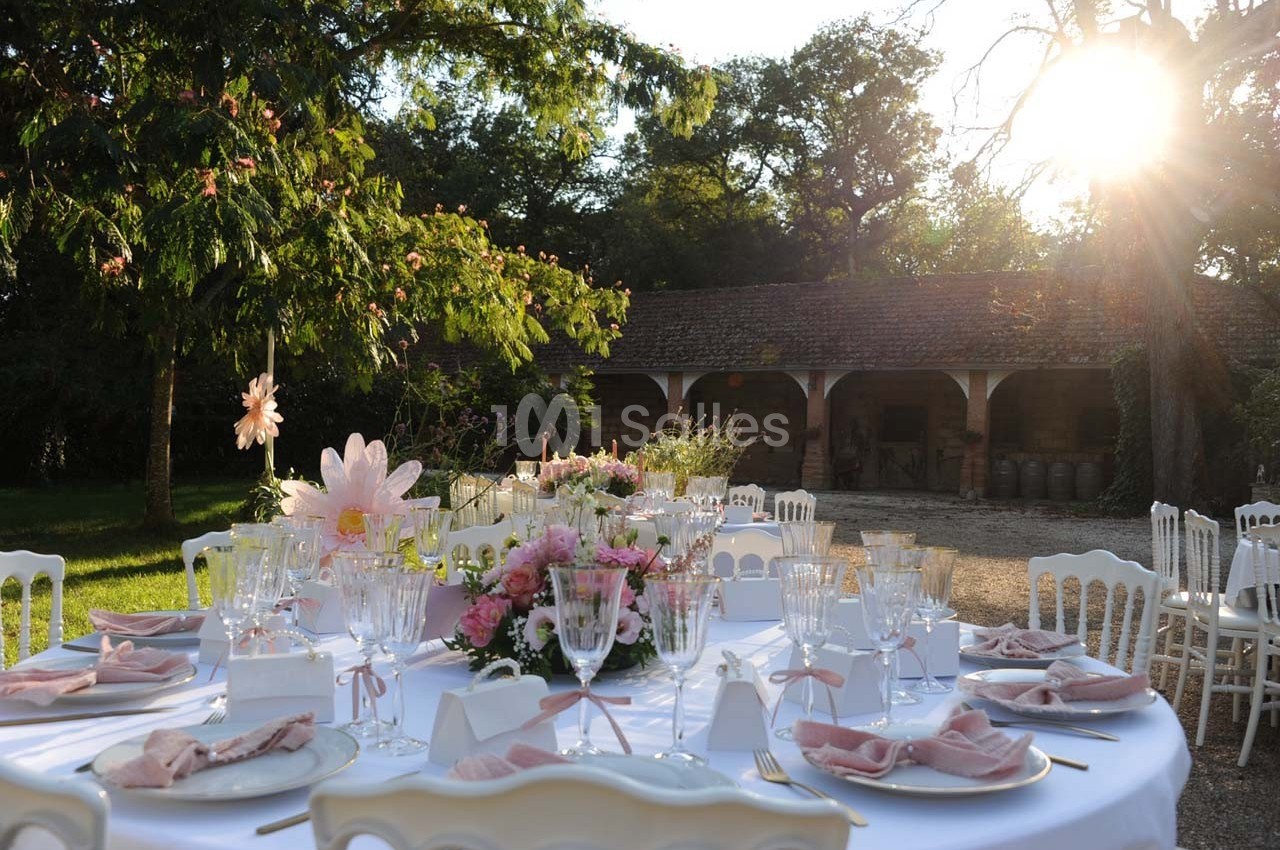 Table dressée en extérieur avec nappes blanches, vaisselle élégante et décorations florales, sous un soleil couchant.