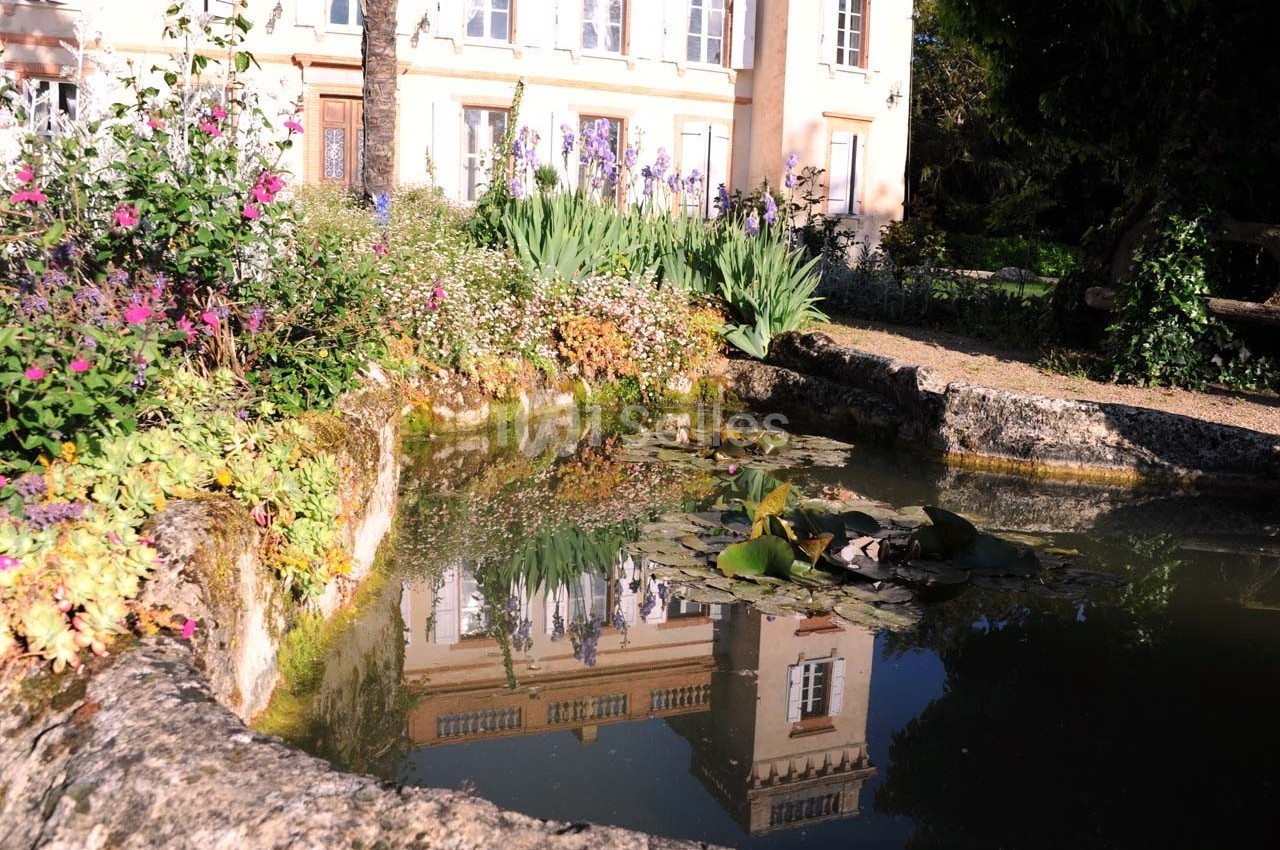 Étang entouré de fleurs et de végétation, reflétant une façade de bâtiment beige sous un ciel ensoleillé.