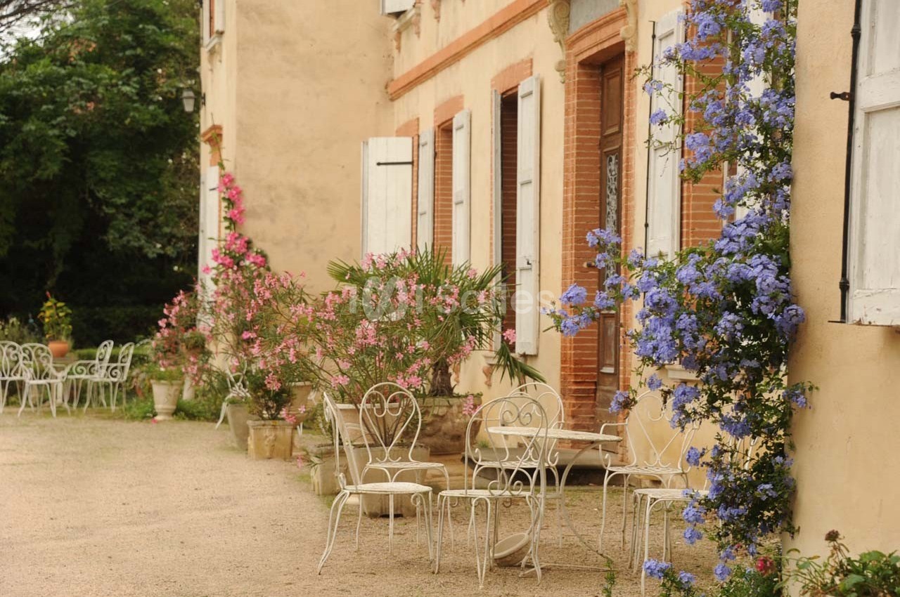 Façade d'une maison ancienne avec volets blancs, mobilier de jardin en fer forgé et plantes fleuries colorées.