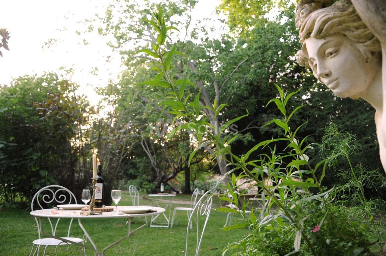 Table de jardin avec bougies et bouteille de vin, entourée de chaises blanches dans un espace vert arboré au crépuscule.