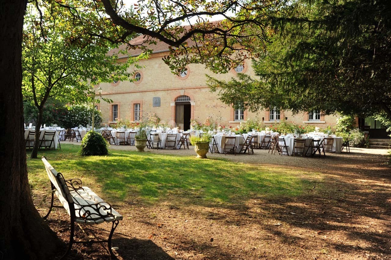 Tables dressées pour un événement en plein air devant un bâtiment en pierre, entouré d'arbres et d'un jardin ombragé.