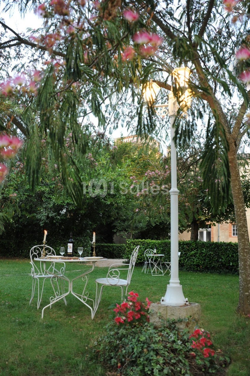 Mobilier de jardin en fer forgé blanc sous un lampadaire allumé, entouré de végétation et de fleurs rouges.