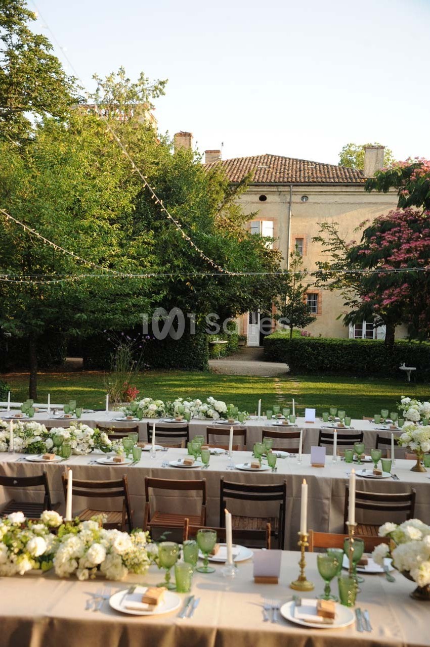 Tables décorées pour un repas en plein air avec nappes beiges, fleurs blanches et bougies, dans un jardin devant un bâtiment…