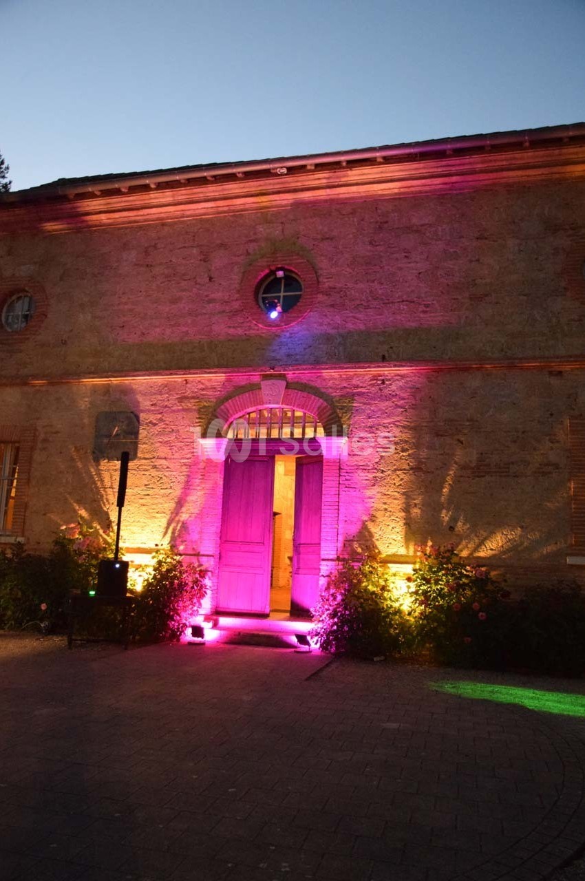 Façade en pierre d'un bâtiment éclairée par des lumières roses et jaunes au crépuscule.