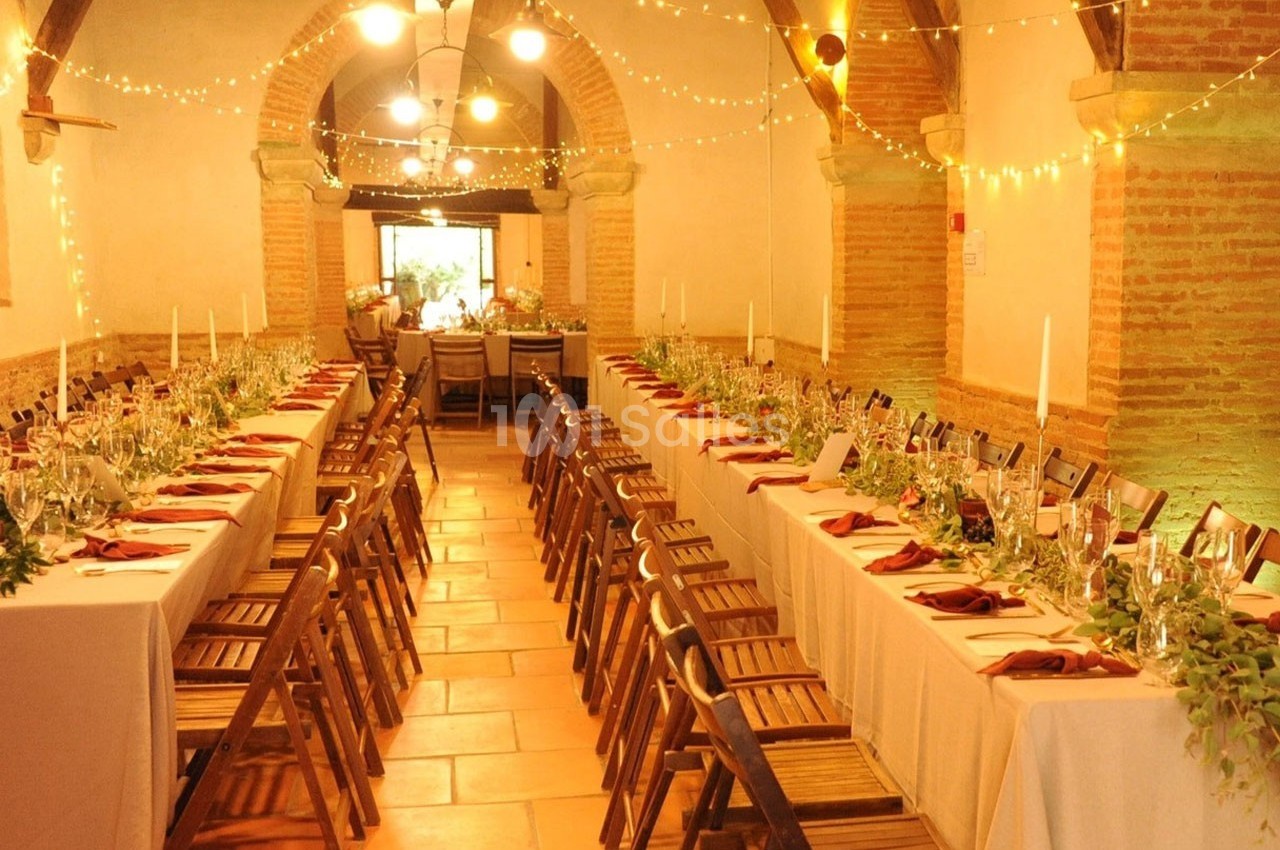 Salle décorée pour un événement avec deux longues tables dressées, chaises en bois et guirlandes lumineuses suspendues.