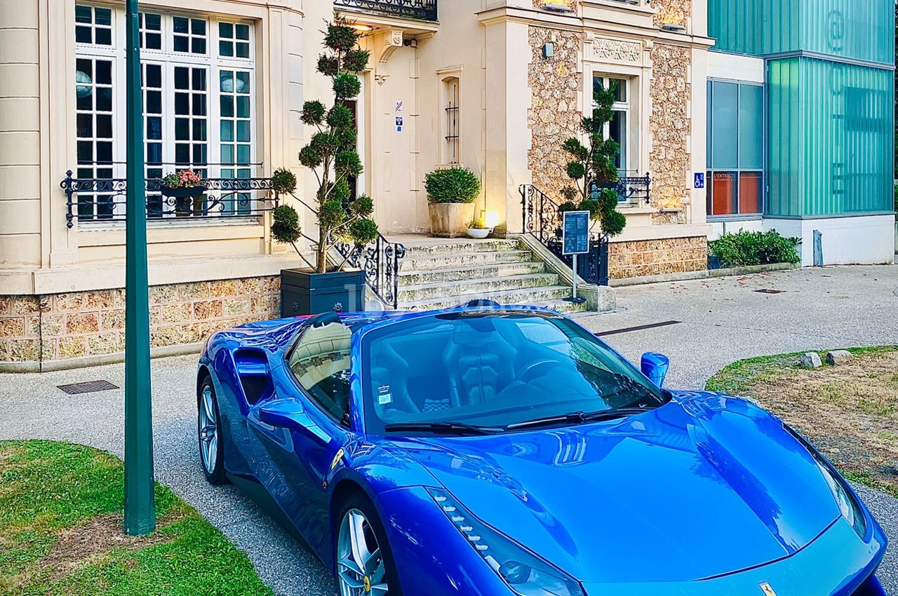 Voiture de sport bleue garée devant une maison de style classique avec des arbustes taillés et un lampadaire.