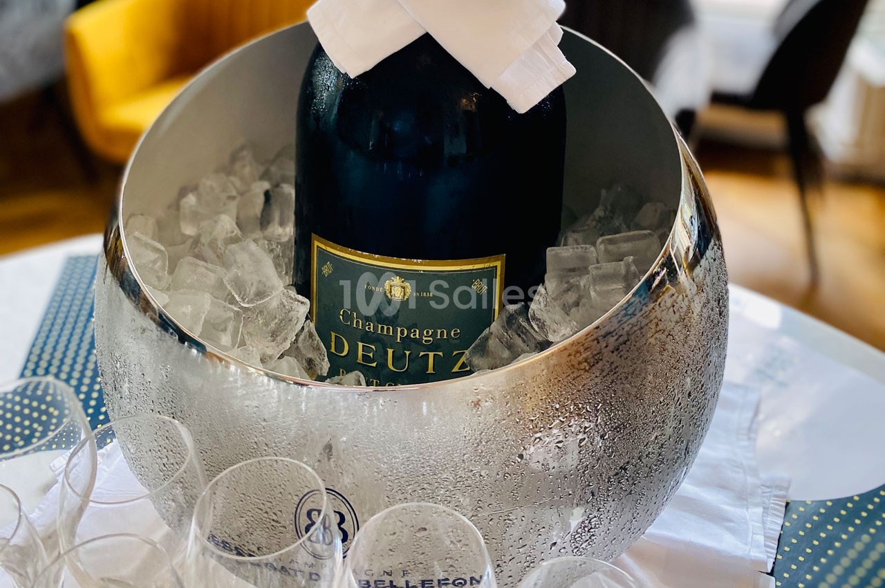 Bouteille de champagne Deutz placée dans un seau argenté rempli de glaçons, entourée de verres vides.