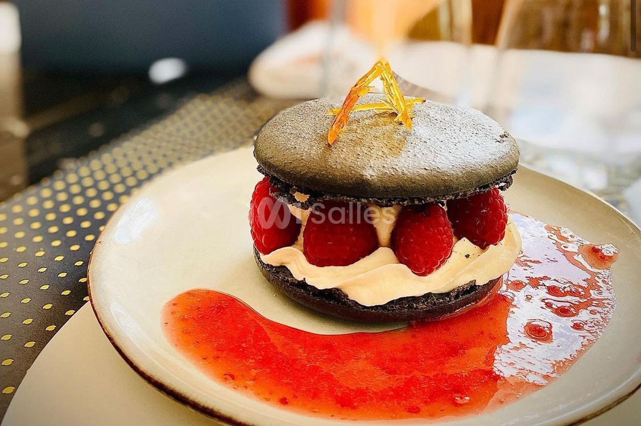 Macaron noir garni de crème et framboises fraîches, accompagné d'un coulis rouge sur une assiette blanche.