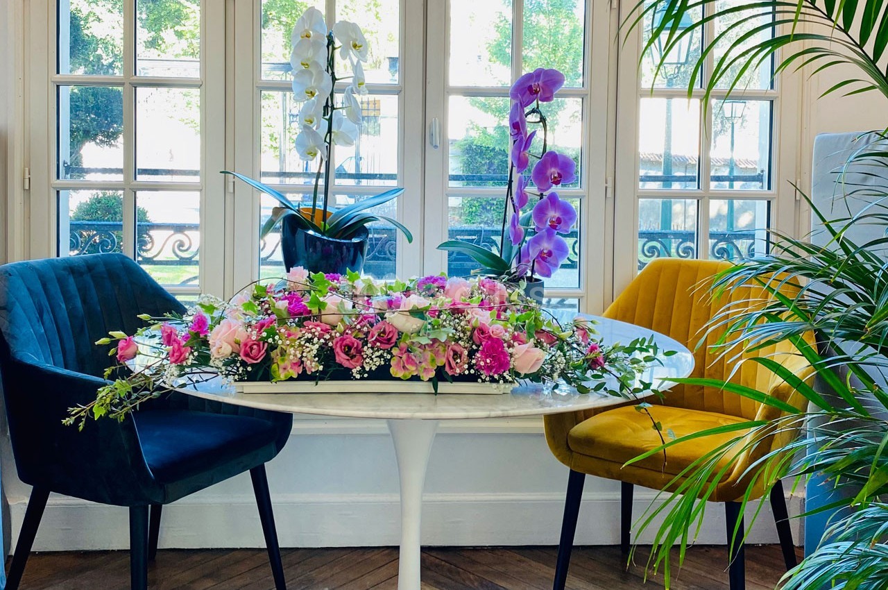 Table ronde décorée de fleurs roses et blanches, entourée de chaises colorées, devant une baie vitrée lumineuse.