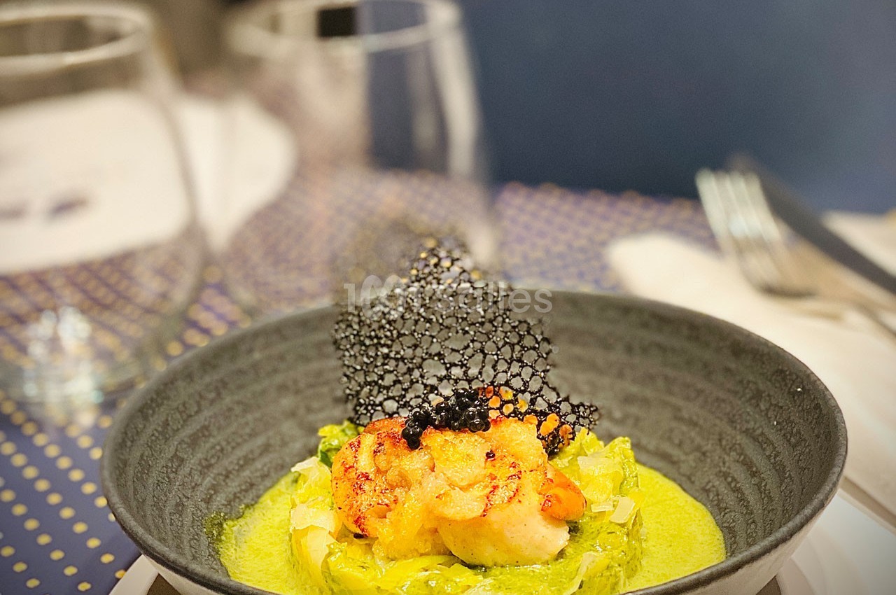 Assiette de crevettes grillées servies sur une purée verte avec une tuile noire décorative, en arrière-plan des verres.