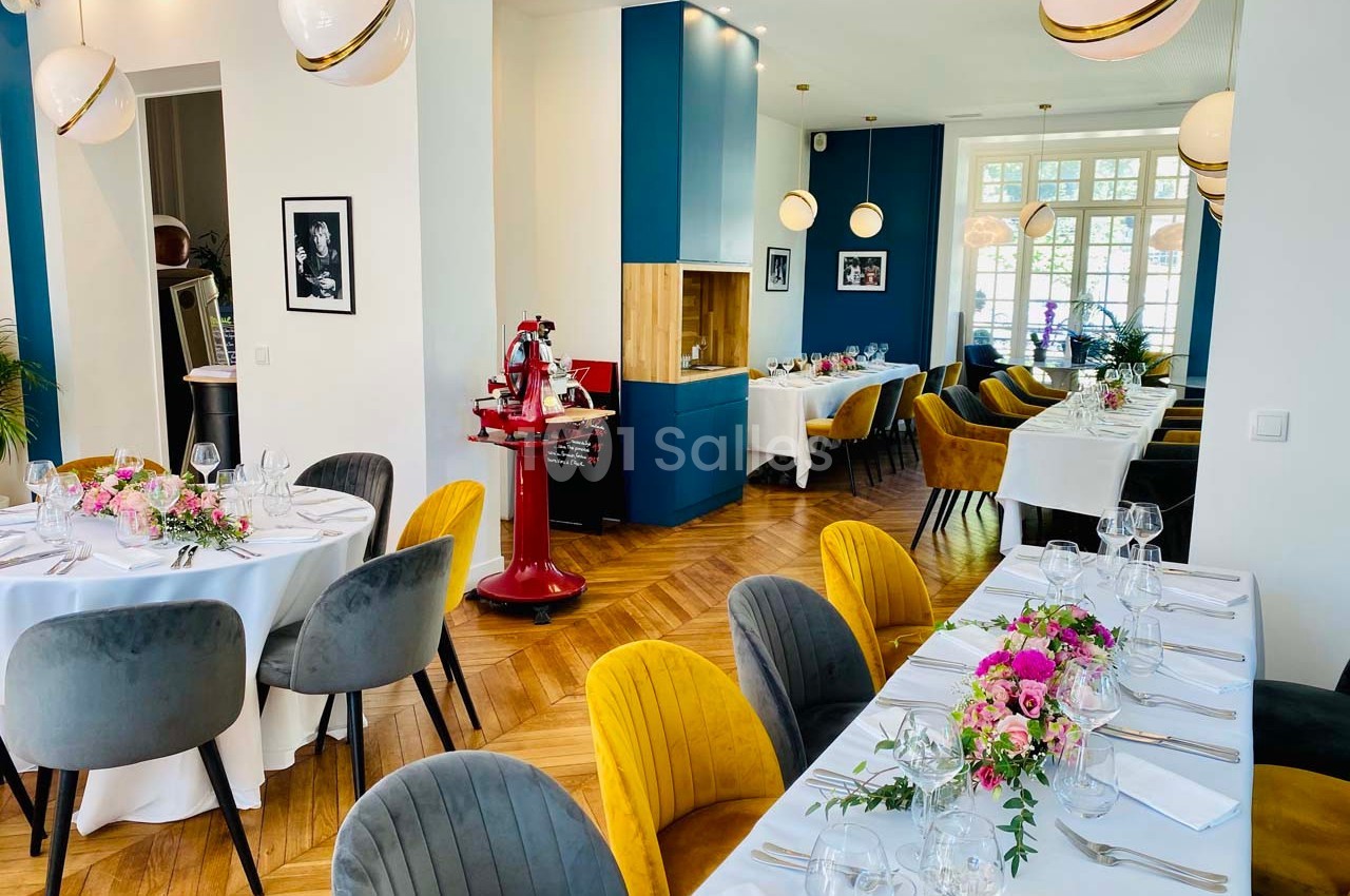 Salle de restaurant lumineuse avec tables dressées, chaises colorées et décorations florales sur un parquet en bois.