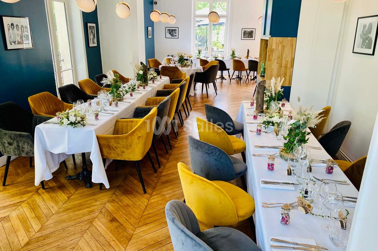 Salle de réception lumineuse avec tables dressées, chaises colorées et décorations florales sur parquet en bois.