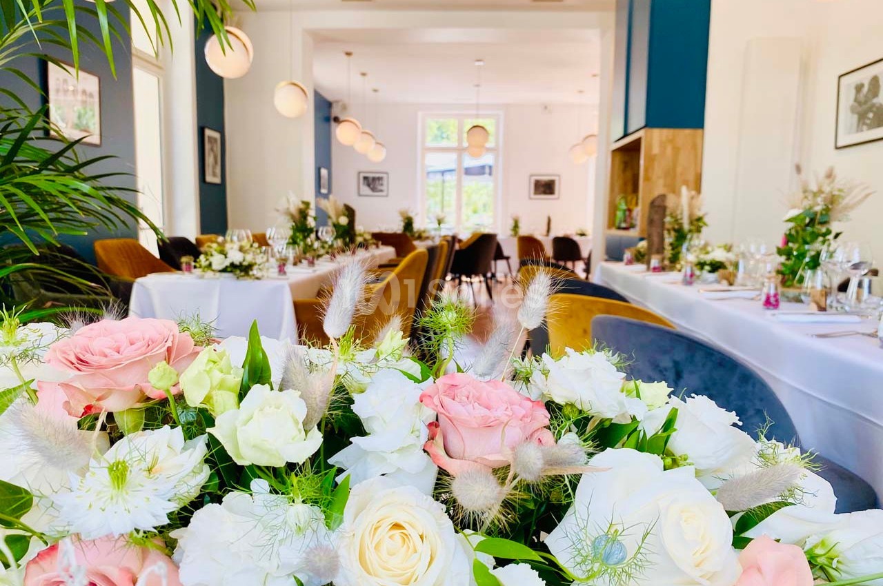 Salle de réception lumineuse avec tables décorées de fleurs blanches et roses, entourées de chaises élégantes.