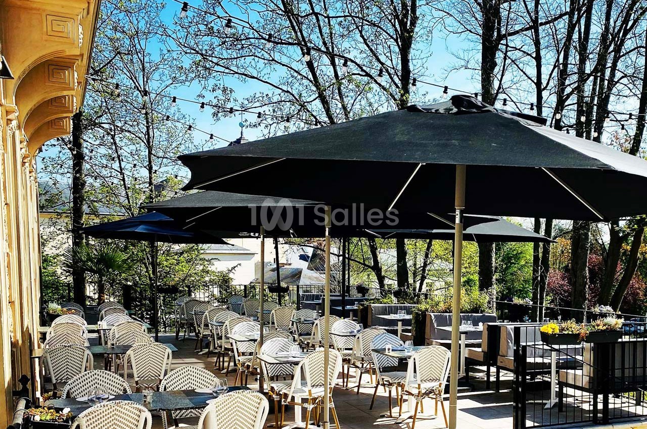 Terrasse ensoleillée avec tables et chaises en osier, ombragée par de grands parasols noirs, entourée d'arbres.