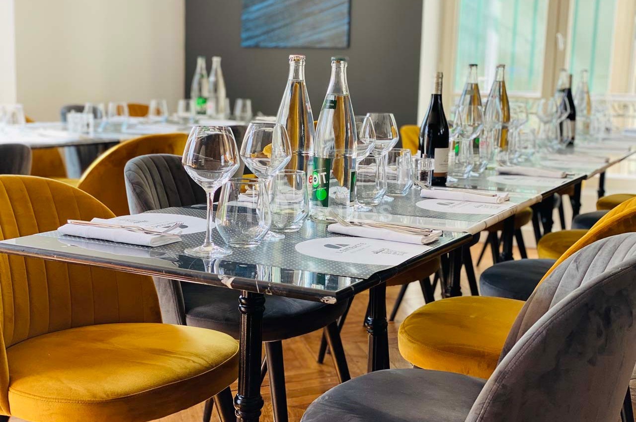 Table dressée avec des verres, bouteilles d'eau et de vin, entourée de chaises jaunes et grises dans une salle lumineuse.