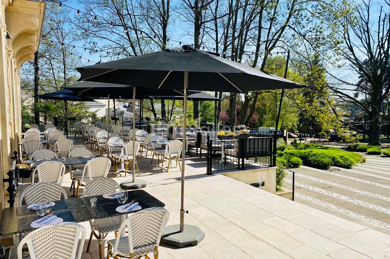 Terrasse d'un restaurant avec tables et chaises en osier, parasols noirs, entourée d'arbres et d'un jardin verdoyant.