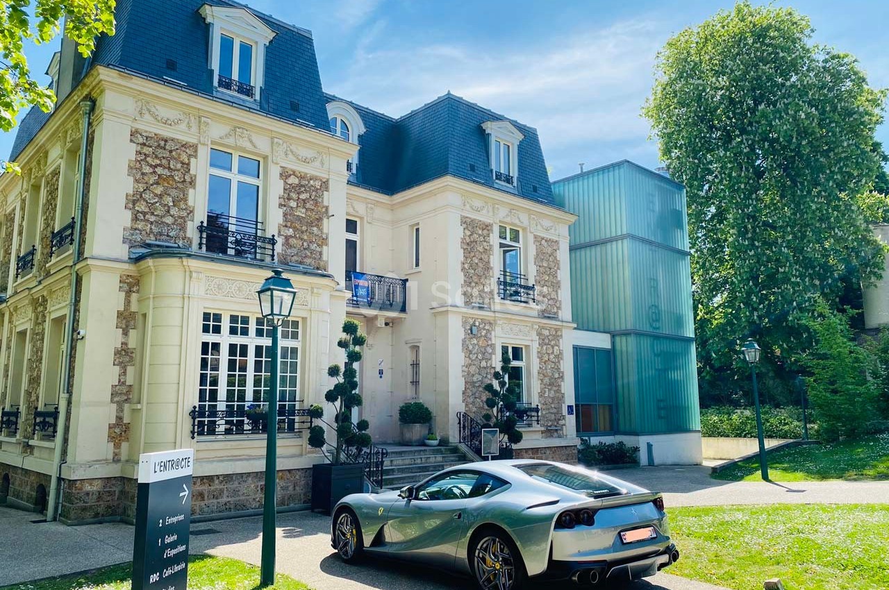 Voiture de sport grise stationnée devant une maison bourgeoise en pierre et un bâtiment moderne en verre.