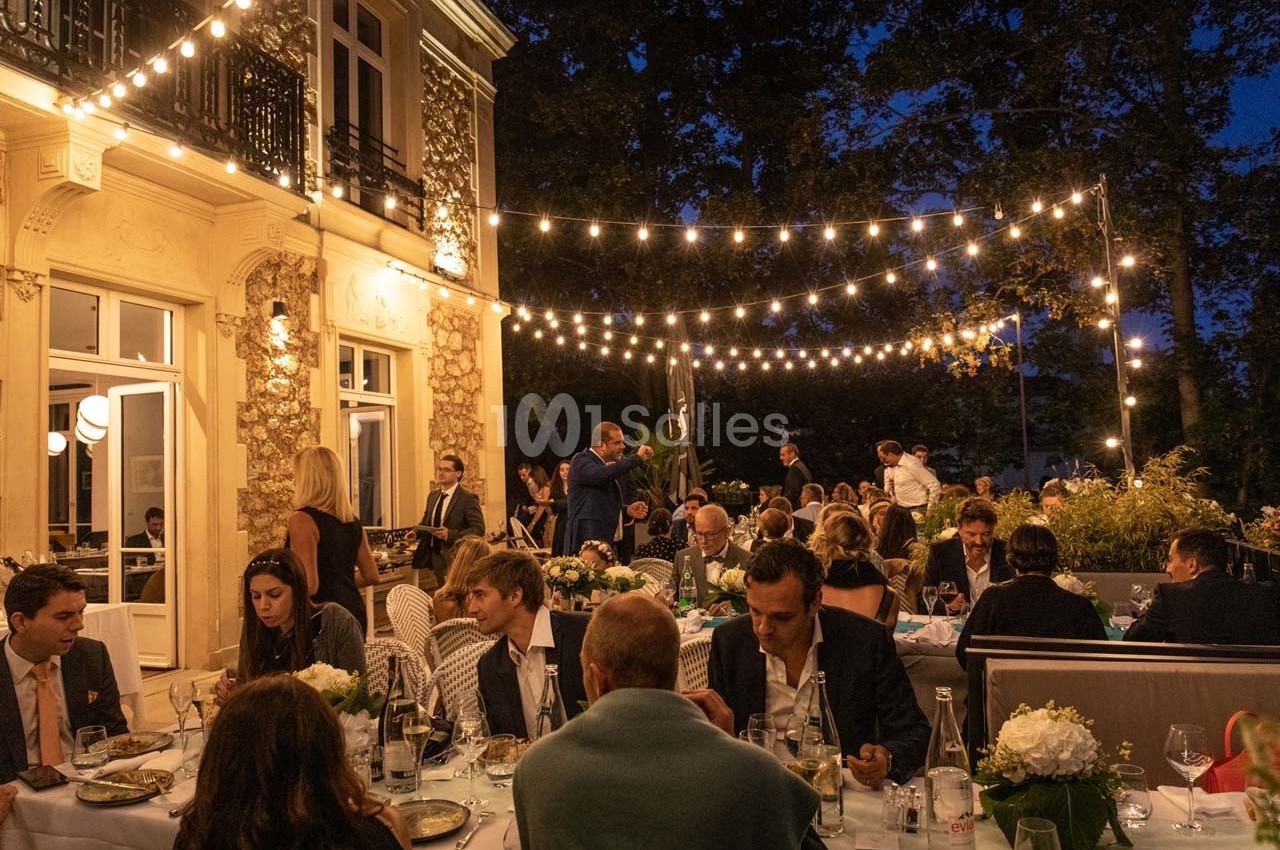 Des convives dînent en soirée sur une terrasse éclairée par des guirlandes lumineuses, près d'un bâtiment élégant.