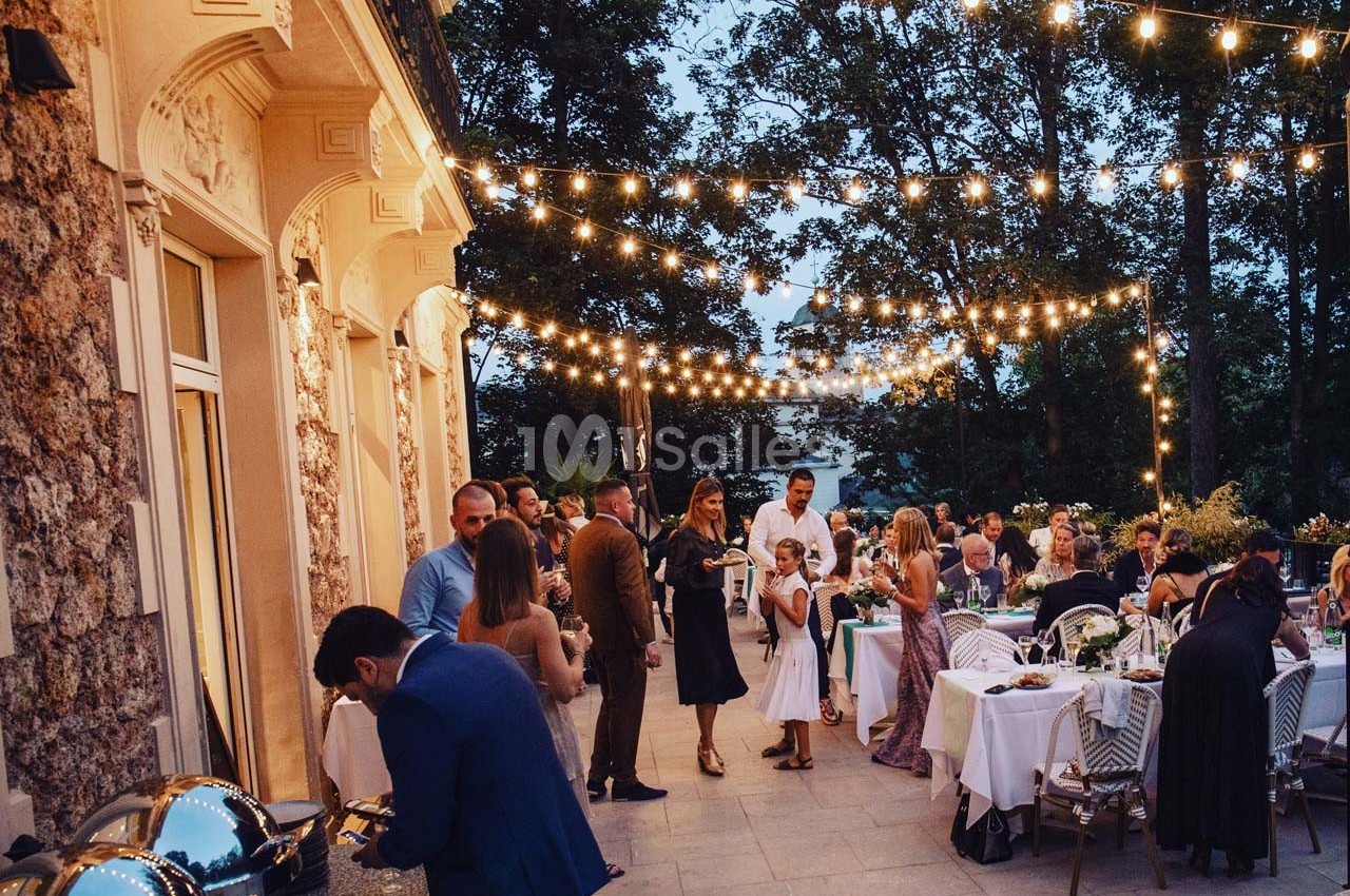 Dîner en plein air avec des invités discutant sous des guirlandes lumineuses, près d'un bâtiment en pierre.