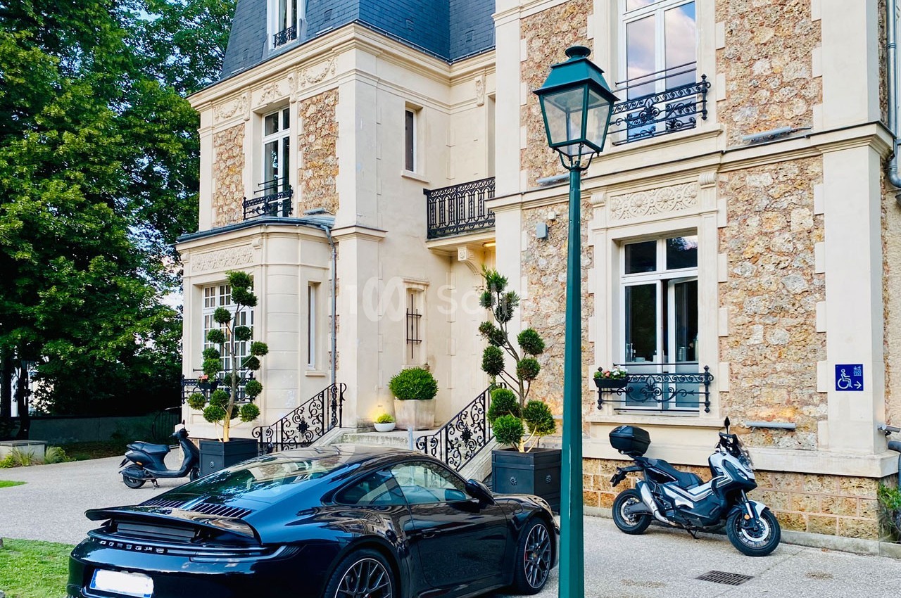 Voiture de sport noire garée devant une maison en pierre avec des escaliers, des plantes et deux scooters à proximité.