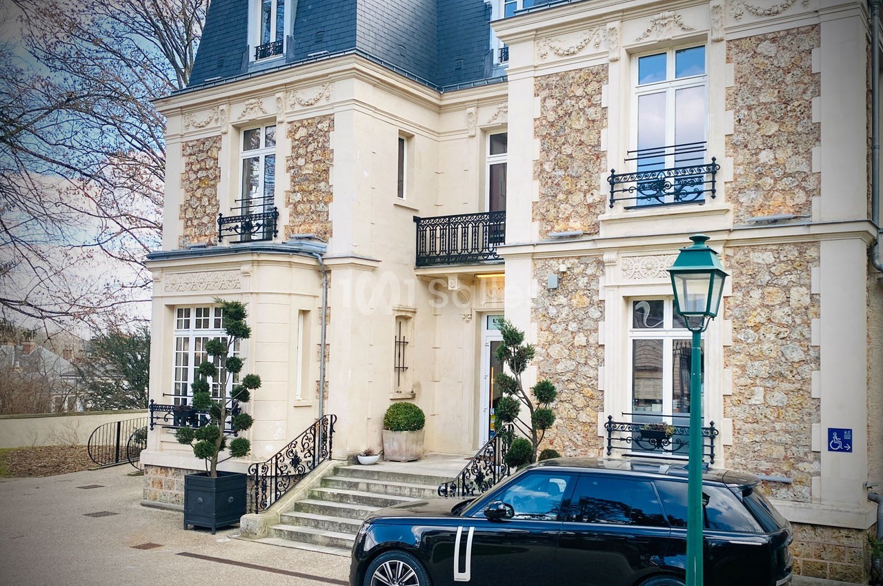 Façade d'une maison bourgeoise en pierre avec un escalier, des plantes taillées et une voiture noire stationnée devant.