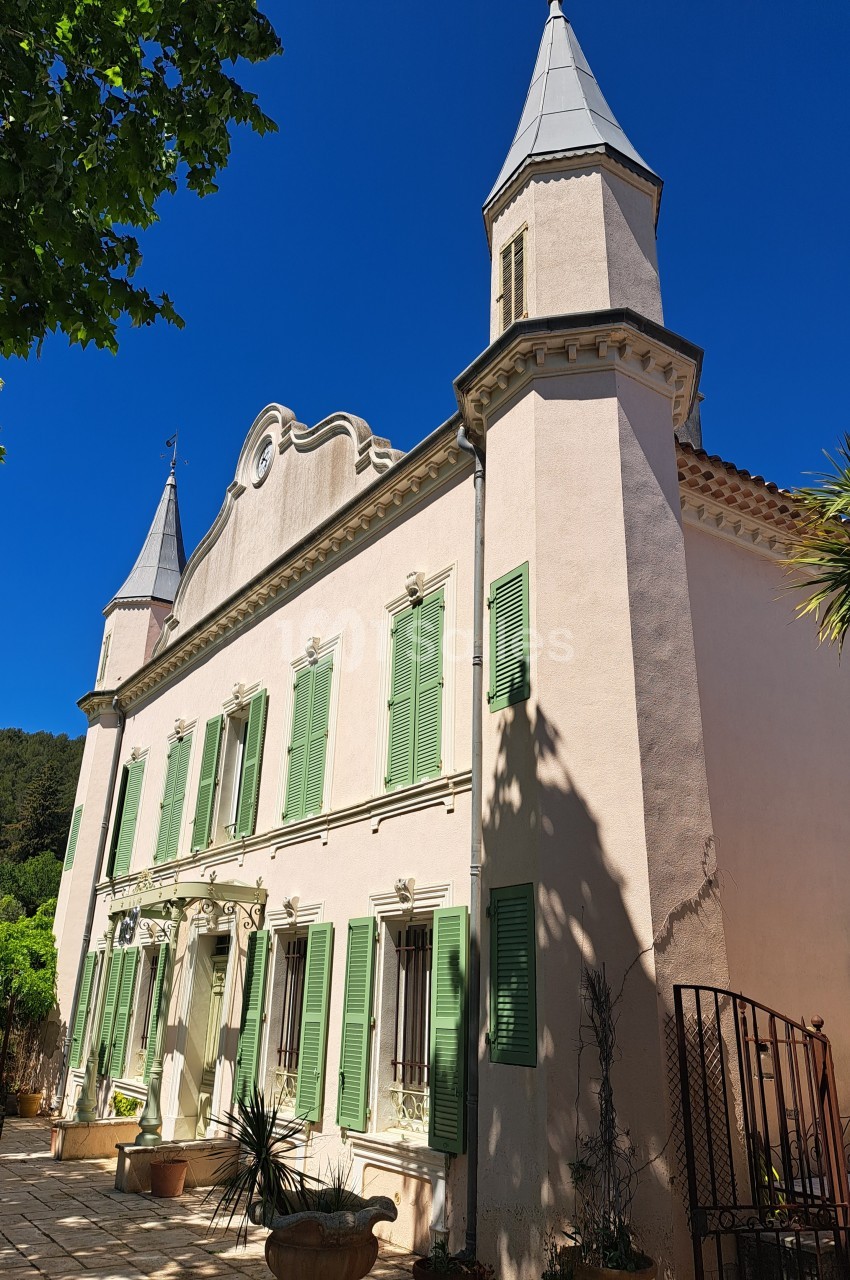 Façade d'une maison ancienne avec volets verts, deux tours et un ciel bleu dégagé en arrière-plan.