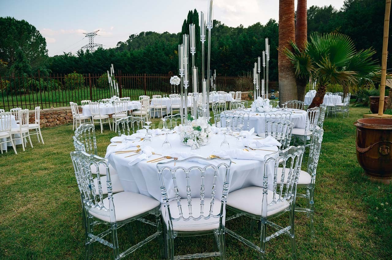 Table ronde élégamment dressée avec nappes blanches, chaises transparentes et centre de table floral, dans un jardin…