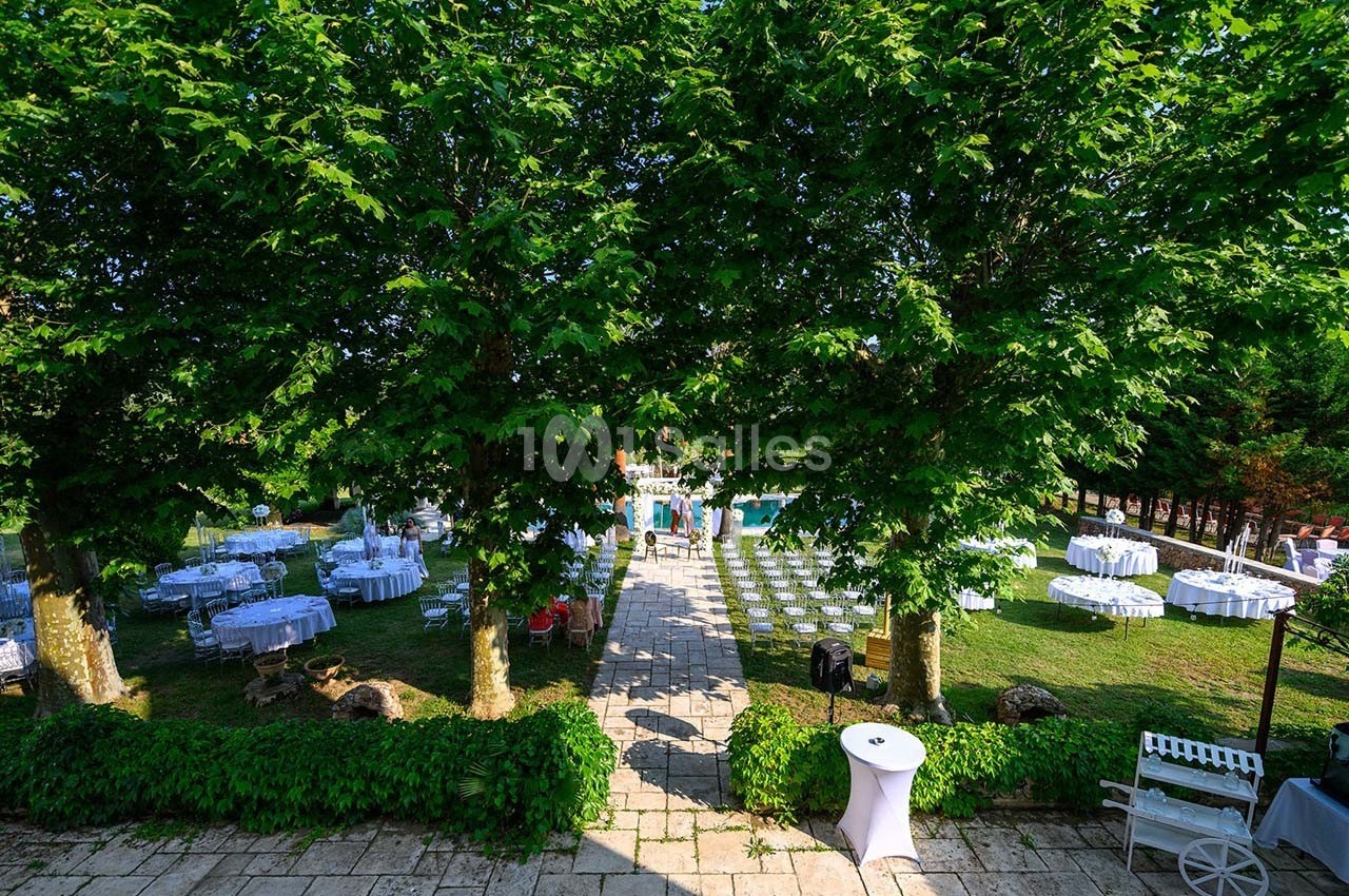 Jardin aménagé pour un événement avec tables rondes dressées, chaises alignées et allée centrale bordée d'arbres.