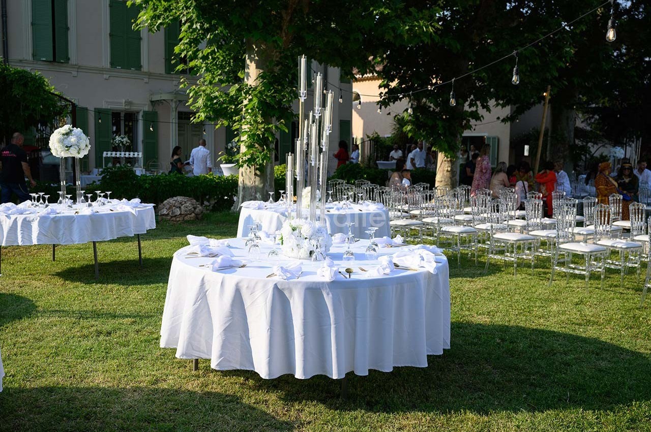 Tables rondes dressées avec nappes blanches dans un jardin, entourées de chaises transparentes et décorées de bougies.