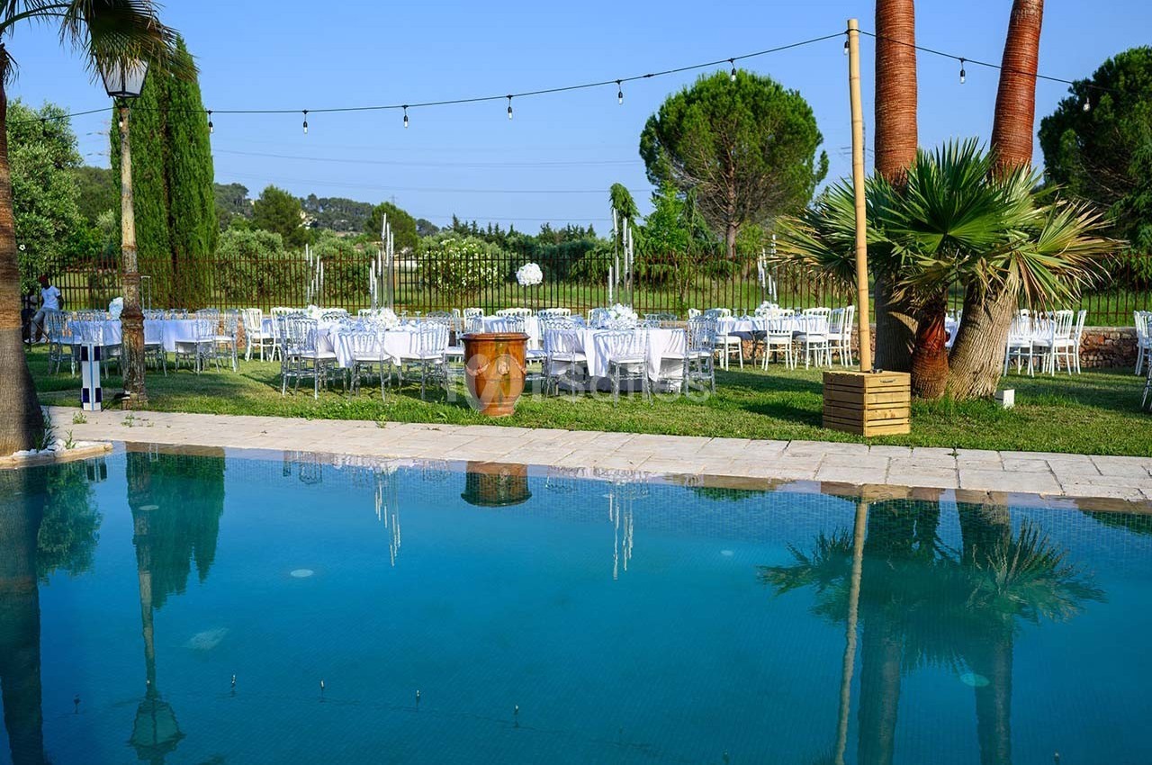Tables dressées en extérieur près d'une piscine, entourées de palmiers et de verdure sous un ciel dégagé.