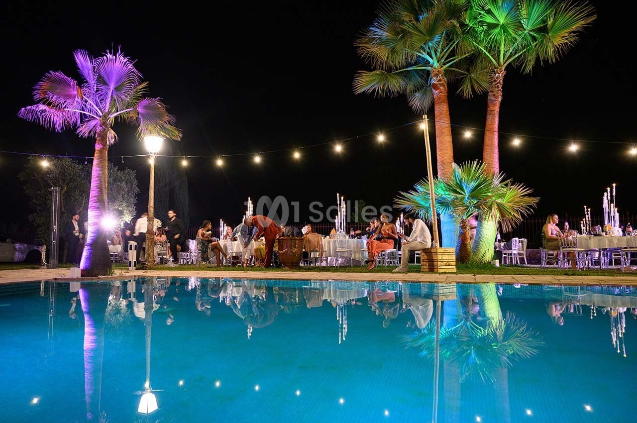 Vue nocturne d'une réception en plein air avec des invités assis près d'une piscine éclairée et entourée de palmiers.