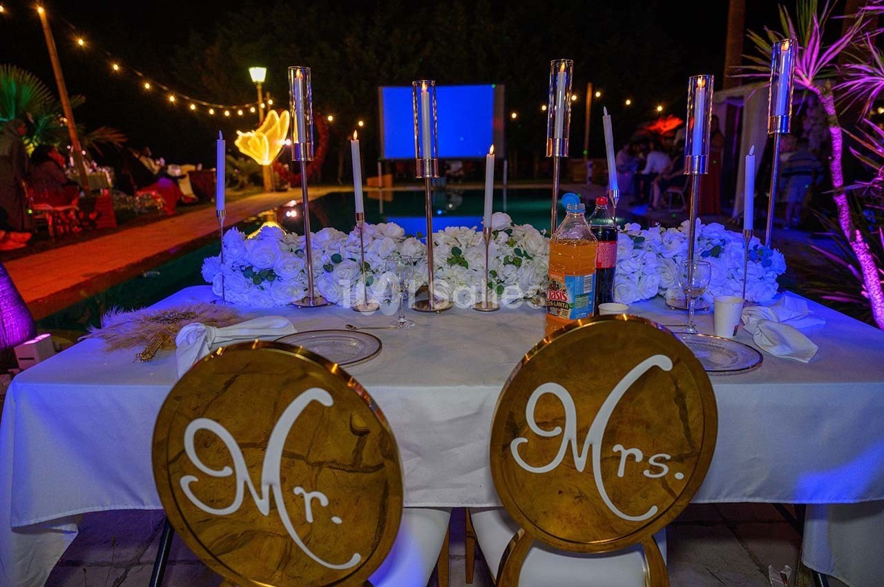 Table décorée pour un mariage avec chandeliers, fleurs blanches et chaises marquées ’Mr.’ et ’Mrs.’ près d'une piscine.