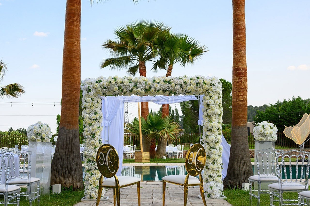 Arche de mariage décorée de fleurs blanches, entourée de palmiers, avec deux chaises dorées devant une piscine.