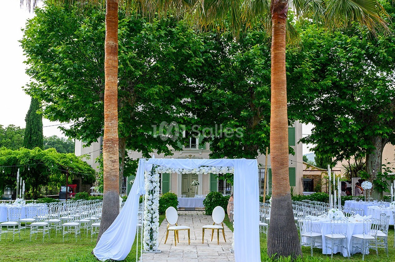 Décoration de mariage en extérieur avec arche blanche, chaises et tables dressées sous des arbres et des palmiers.