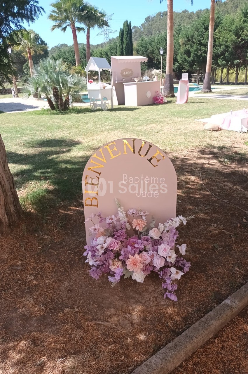 Panneau ’Bienvenue Baptême Jade’ décoré de fleurs roses et violettes, installé dans un jardin ensoleillé.