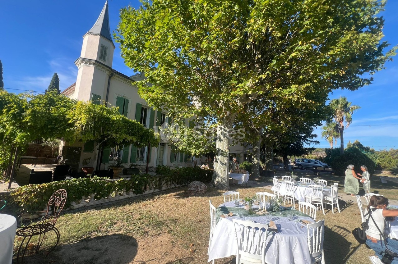 Tables dressées en extérieur sous des arbres, près d'une grande maison avec des volets verts par une journée ensoleillée.