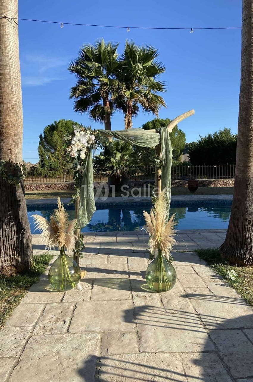 Arche de mariage décorée de tissu vert, fleurs et pampas, installée entre deux palmiers près d'une piscine.
