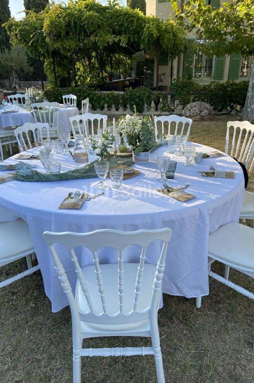 Table ronde dressée en extérieur avec nappe blanche, chaises blanches et décoration florale, sous des arbres.